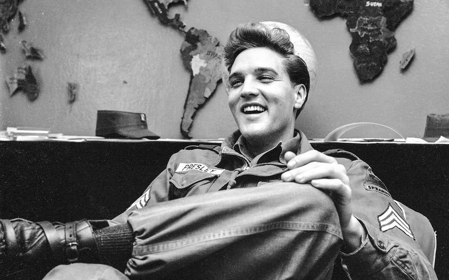 Elvis Presley wears an Army uniform and leans back, crosses one leg over the other, and smiles. 