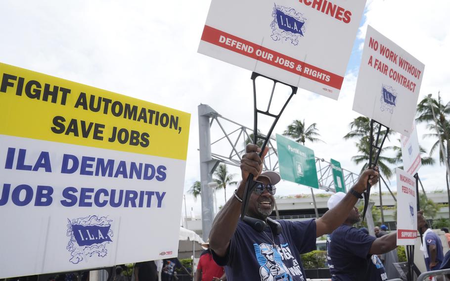 Striking dockworkers hold up signs while standing on a picket line.