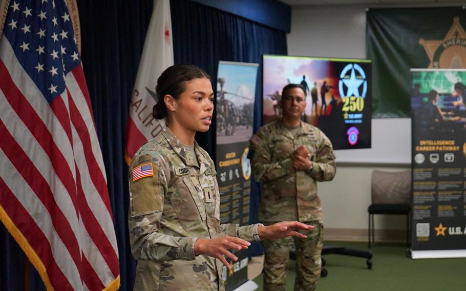 A soldier speaks with her hands out in front of the USA flag