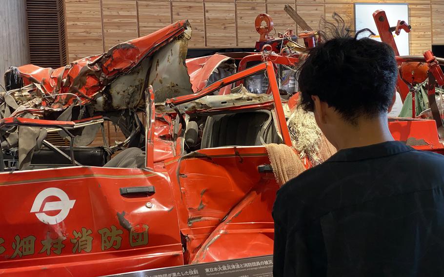 A man with dark hair wearing a dark shirt, seen from behind, views a damaged red car as part of a museum exhibit.