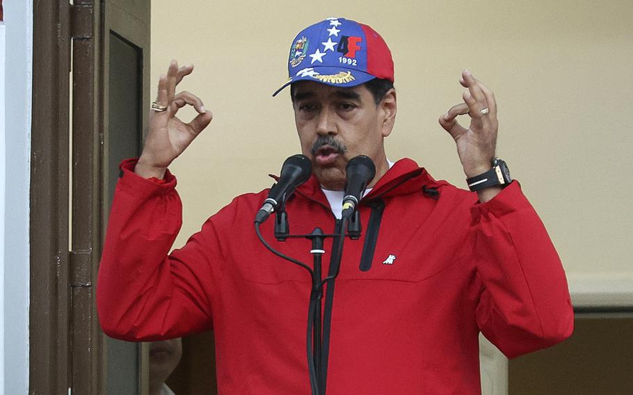 Nicolas Maduro speaks before a microphone in a red jacket, with both hands raised up in gesture.