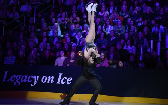 With the crowd in the background a male skater lifts a female skater, upside down, into the air.