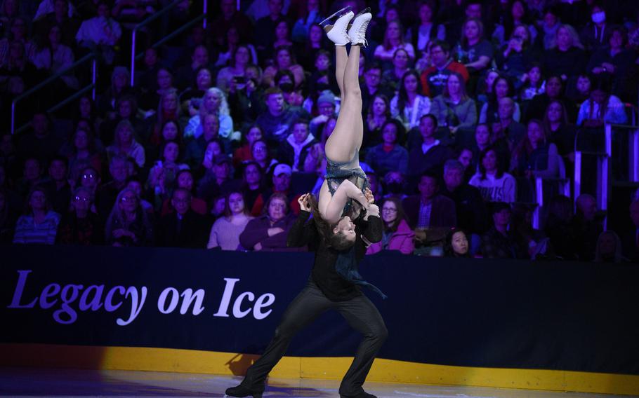 With the crowd in the background a male skater lifts a female skater, upside down, into the air.