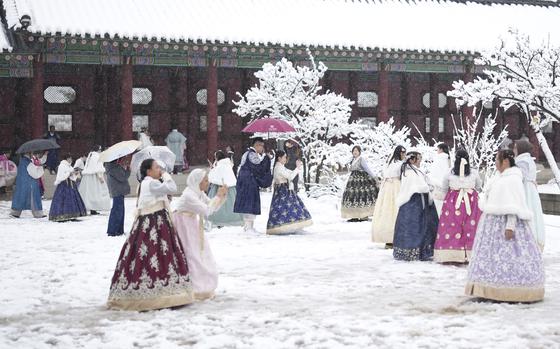 Young women dressed in colorful hanboks take pictures in the snow on the grounds of a red palace.