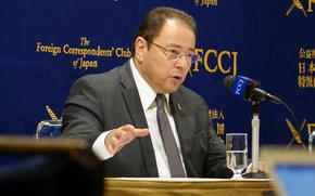 The Ukrainian Ambassador to Japan, wearing a suit and glasses, sits at a table and speaks into a microphone.