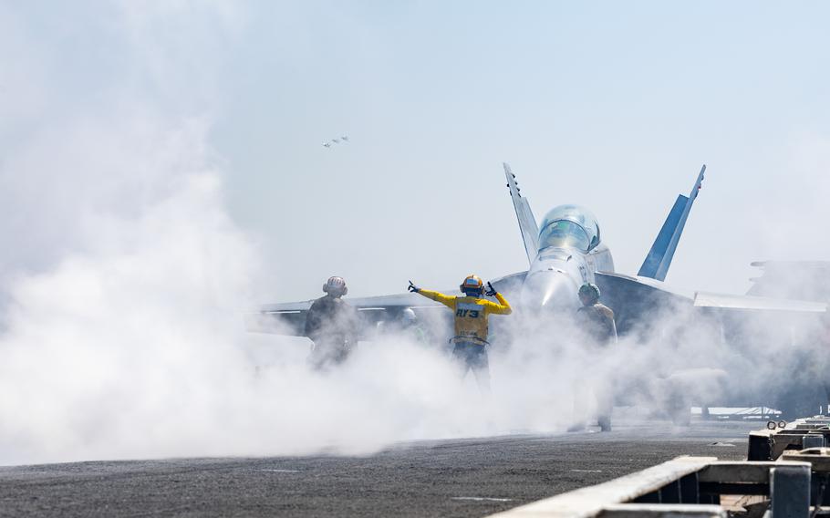 Two crewmembers on the deck of an aircraft carrier direct a jet for takeoff.