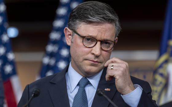 Mike Johnson, wearing a dark suit and glasses, speaks into microphones while standing at a podium in front of U.S. flags.