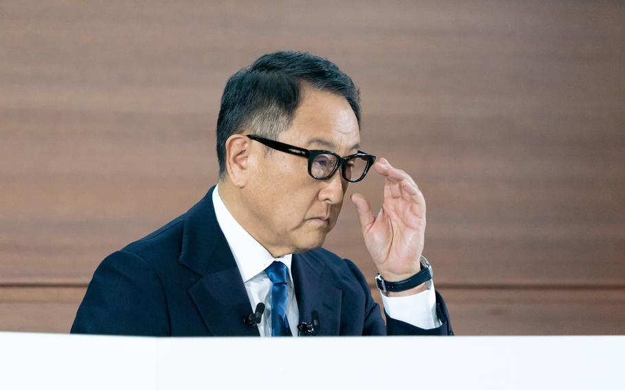 A man in a suit touches his glasses with one hand while most of his body is obscured by a white table or other surface.