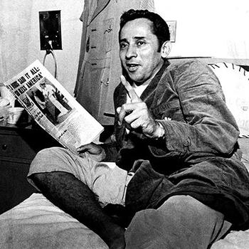 Black and white photo of a man in 1970s-era military attire lying on a hospital bed and holding a newspaper.