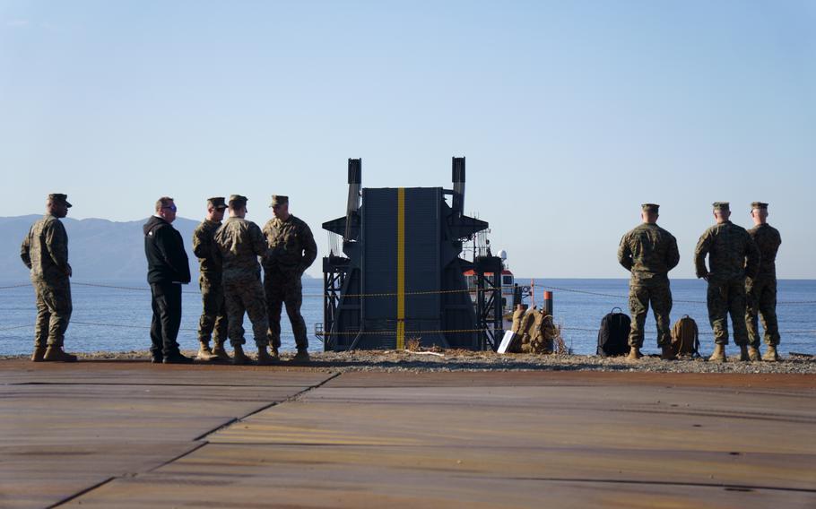 Marines in camouflage uniforms stand on a loading dock area with a bay in the background.