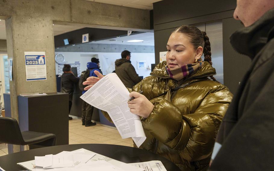 A woman looks at a ballot with other people in the background lining up at a clerk counter.