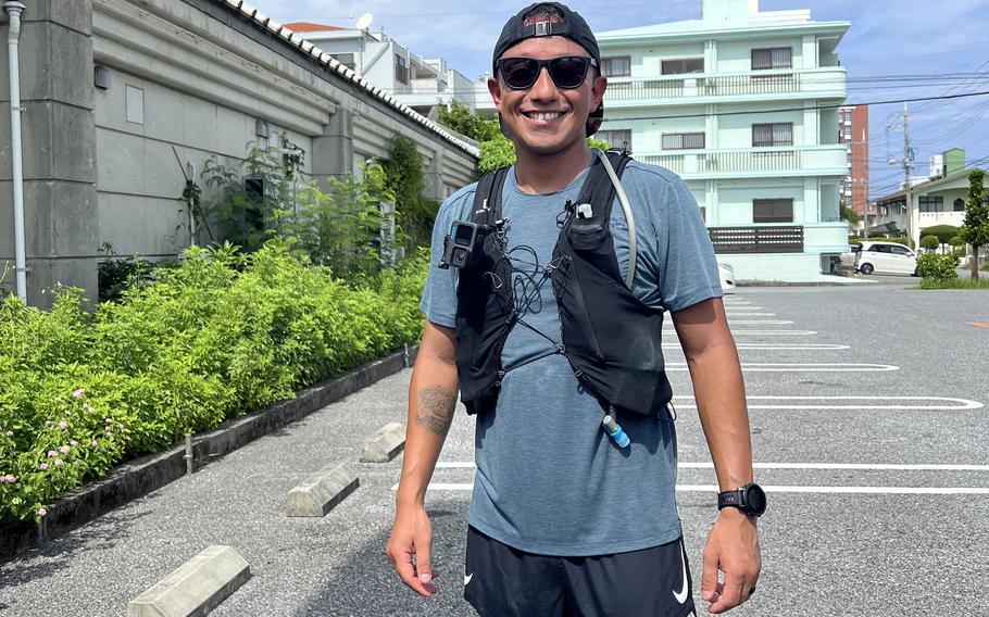 A Marine wearing sunglasses, a backwards baseball cap, a blue T-shirt, a black hydration pack vest and navy blue shorts grins.