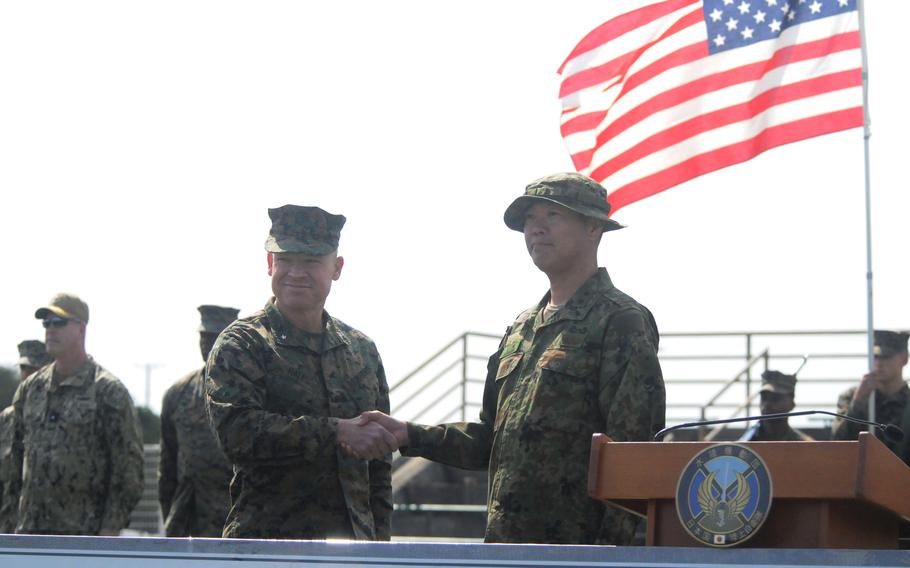 A U.S. brigadier general shakes hands with a Japanese major general with service members and an American flag in the background.