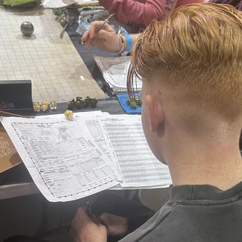 A man, seen from behind, looks at a Dungeons and Dragons character sheet on a table next to dice and a map board.