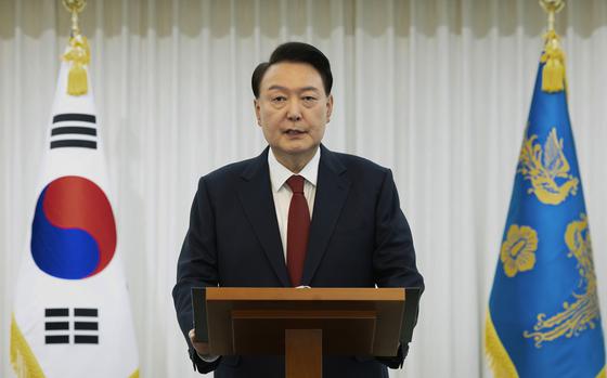 A man in a black suit and red tie speaks at a podium in front of a Korean flag.