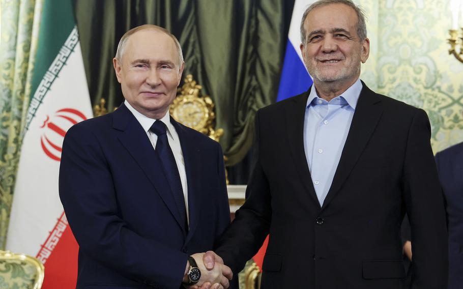 Two men in suits, one blue and one black, shake hands and smile for the camera.