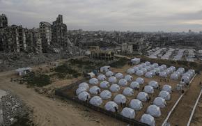 Rows of white tents in a walled enclosure, seen from above, sit next to a skyline of destroyed buildings.