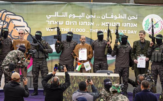 Masked men holding rifles in the air next to restrained hostages by a press conference table.