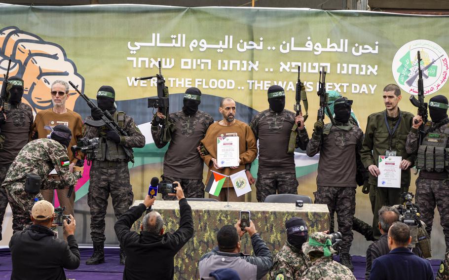 Masked men holding rifles in the air next to restrained hostages by a press conference table.