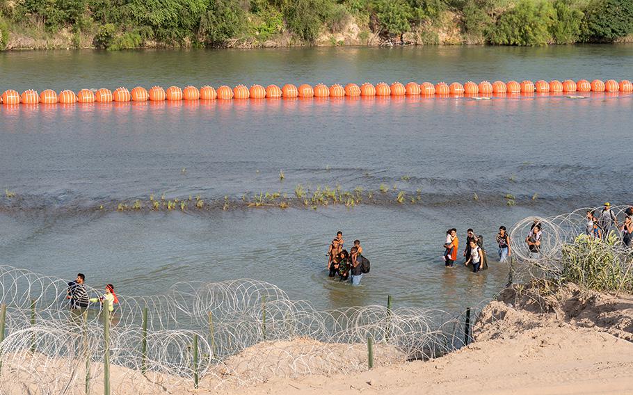 Migrants walk by a string of buoys placed on the water along the Rio Grande border with Mexico in Eagle Pass, Texas, on July 16, 2023. The buoy installation is part of an operation Texas is pursuing to secure its borders, but activists and some legislators say Governor Greg Abbott is exceeding his authority. 