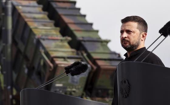 Ukrainian President Volodymyr Zelenskyy stands at a microphone on a podium in front of a missile defense system.