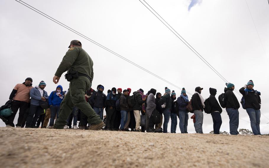 A group of migrants waits in a line as a border patrol agent walks past.