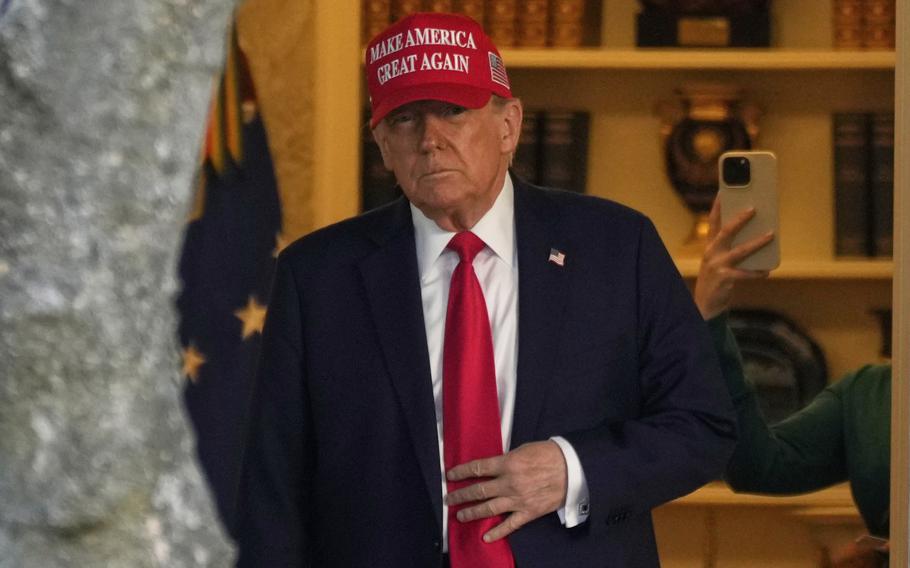 Donald Trump, wearing a dark suit and red “Make America Great Again” hat stands in a doorway of the Oval Office.