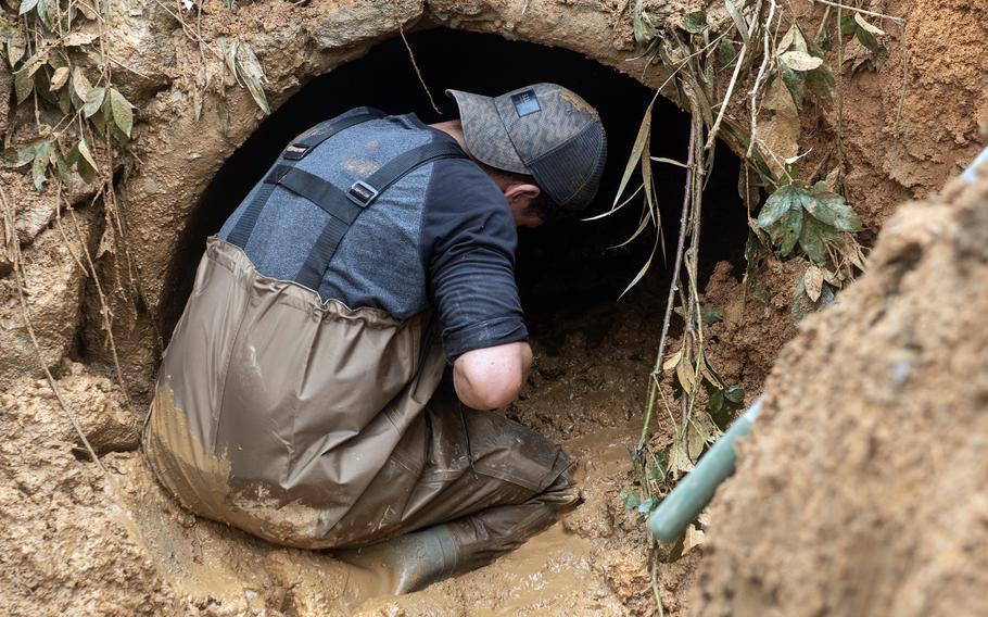 A man in waders kneels in the mud in front of the entrance to a drainage pipe while on a recovery mission.