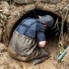 A man in waders kneels in the mud in front of the entrance to a drainage pipe while on a recovery mission.