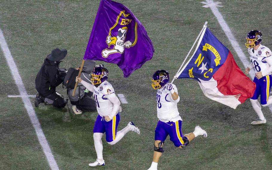 East Carolina football players carrying flags run across the football field.