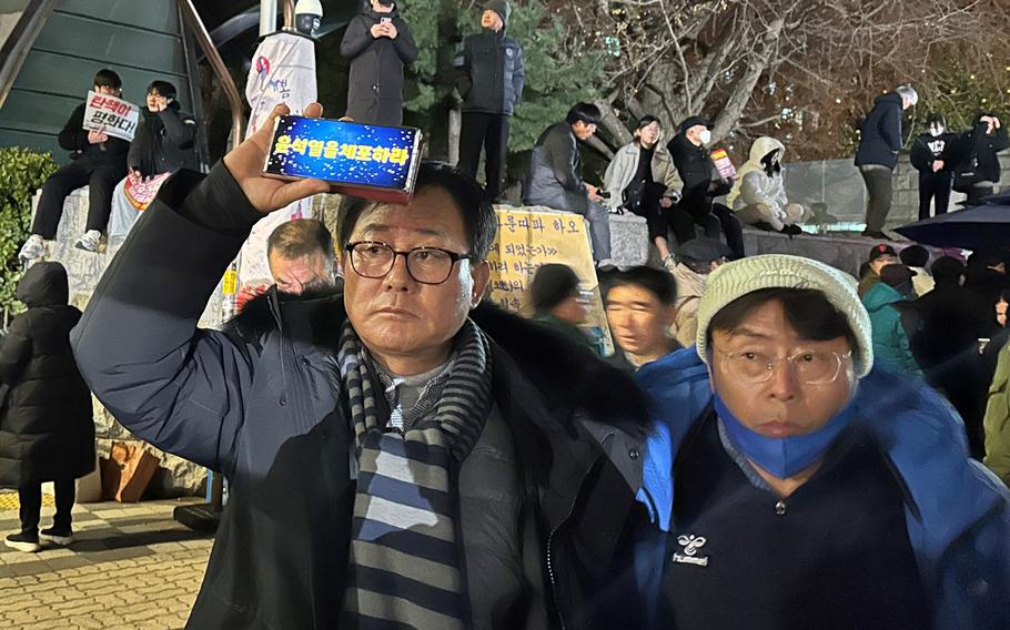 A man holds a phone that reads “Arrest Yoon Suk Yeol” as protesters stand in the background.