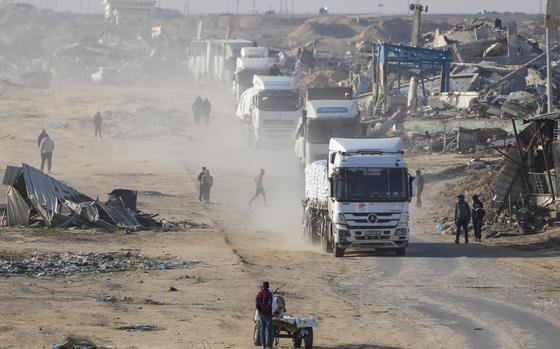 A convoy of white trucks drive through a path with rubble on either side of it.