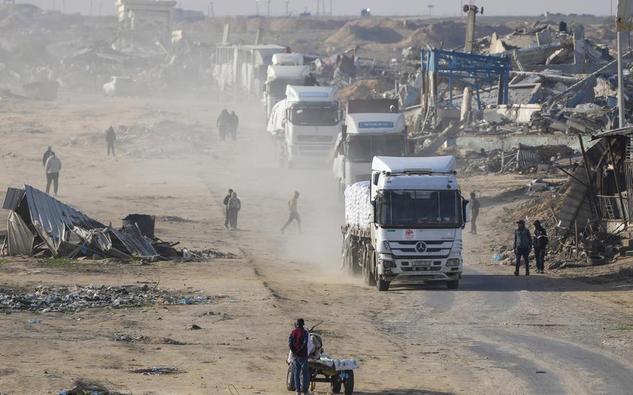 A convoy of white trucks drive through a path with rubble on either side of it.