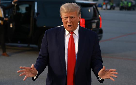 Donald Trump speaks with his arms outstretched to the sides on the tarmac of an airport while a dark SUV waits in the background.