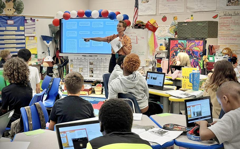 Teacher points to a large screen as she instructs students sitting in front of computers.