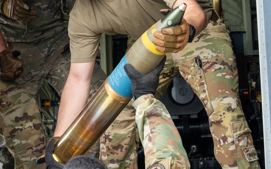 A soldier in camouflage hands a large 105mm shell to another while loading an aircraft.