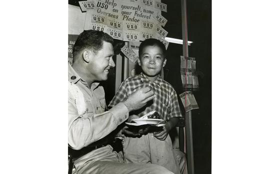 A young Korean boy stands next to a seated U.S. service member holding a plate with a piece of cake.