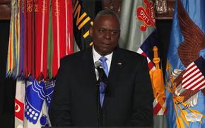 Secretary of Defense Lloyd J. Austin III speaks into a microphone with many different colorful military flags behind him.