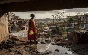 A girl in the foreground holds a bucket, while the wreckage of cyclone-struck slums is in the background.