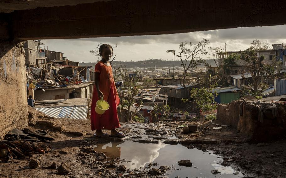 A girl in the foreground holds a bucket, while the wreckage of cyclone-struck slums is in the background.