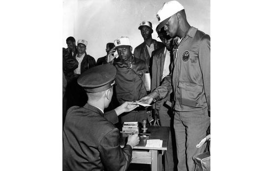 A black-and-white image of an American athlete in a track suit handing a form to a uniformed official seated at a table.