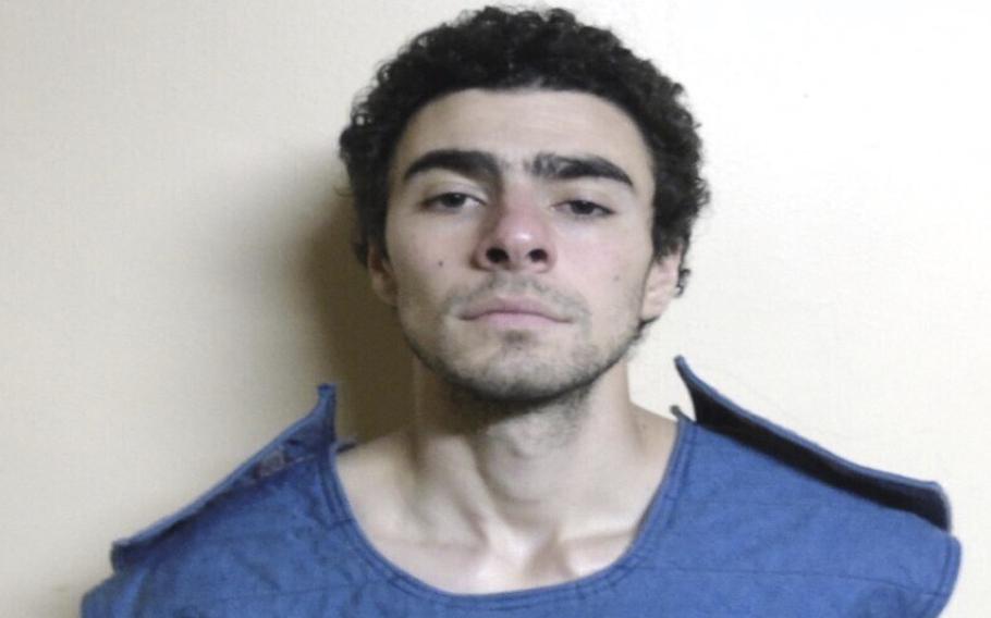 A police booking photo of a man with dark, curly hair wearing a blue shirt and standing in front of a white wall.