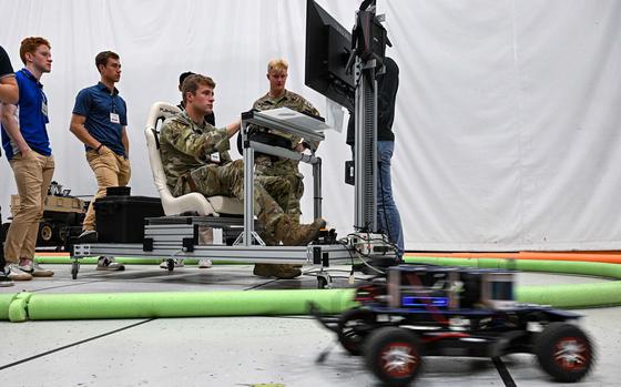 A service member in uniform sits at a keyboard in front of a monitor as cadets look on. 