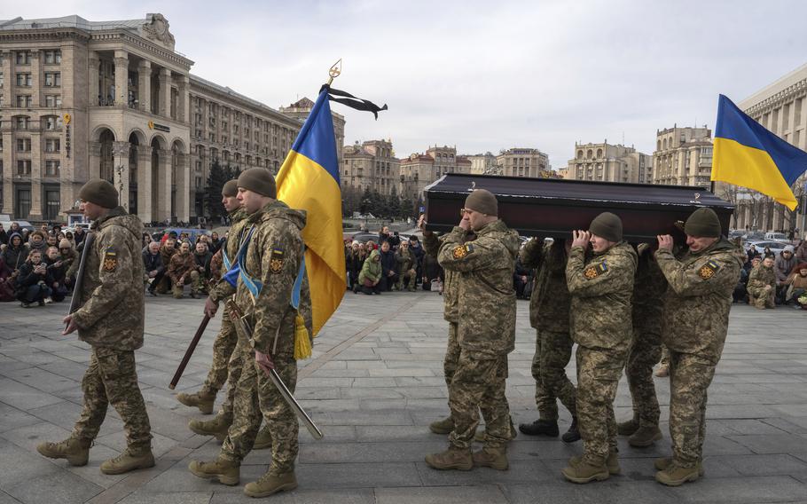 Ukrainian servicemen carry their national flag and a casket before an audience in a public square.