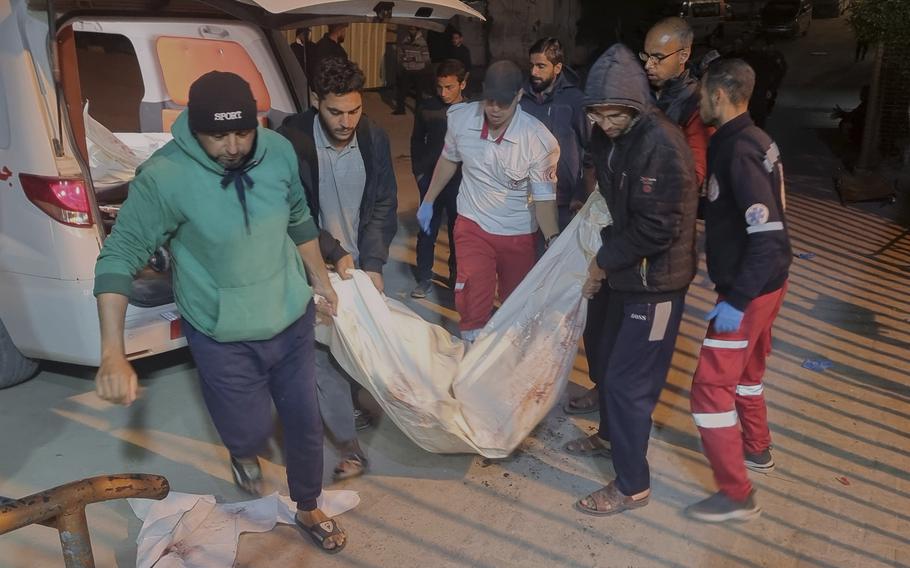 A group of men carry a body wrapped in a sheet out of the back of an ambulance.