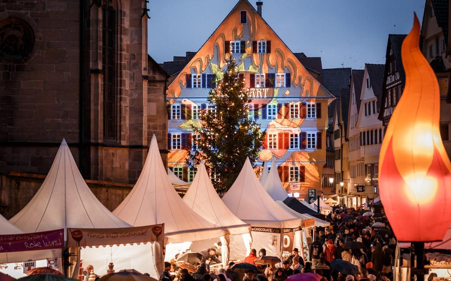 Festive, brightly lit market stalls attract onlookers with a Christmas tree and blue-painted Bavarian house with red shutters in the background. 