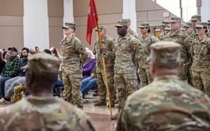 A group of sldiers stand in formation, seen past the soulders of two soldiers seated in front of them.