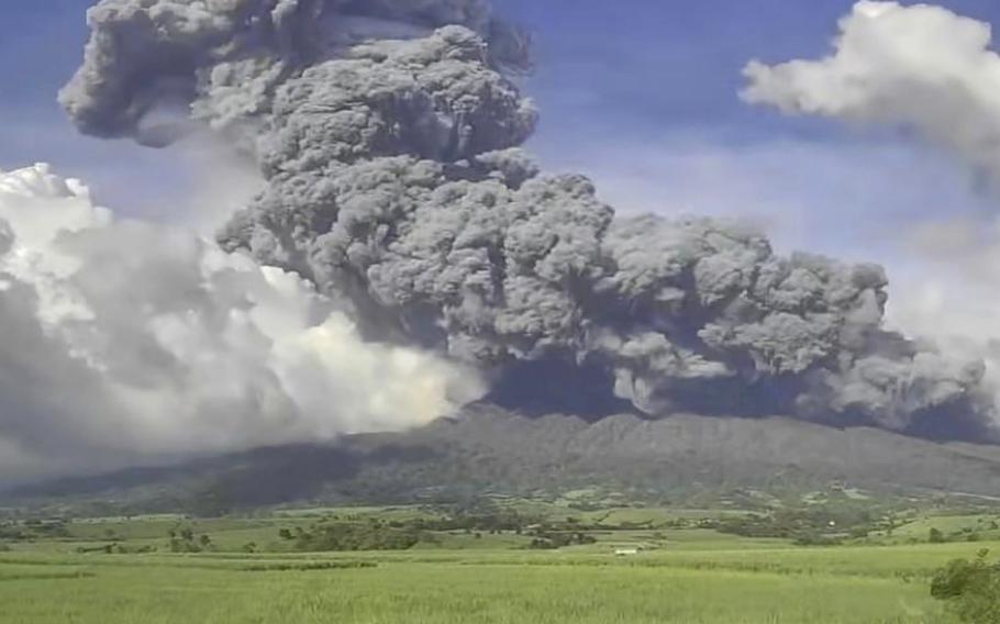 A volcano above green grass belches gray smoke into a bright blue sky.