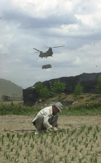 A farmer crouches over his field while a helicopter flies in the background.