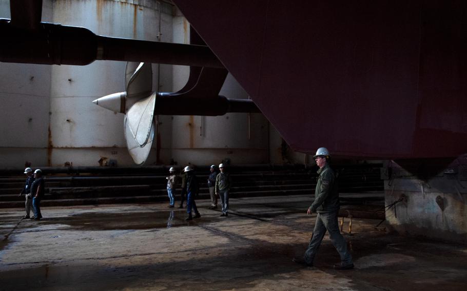 Several men in hard hats walk around the large propeller of an aircraft carrier as it sits in dry dock.